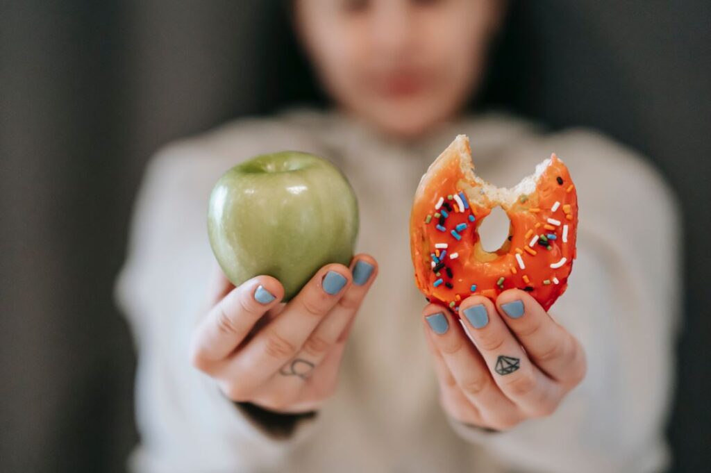 Mujer sosteniendo a cámara una manzana verde en una mano y una dona en la otra.