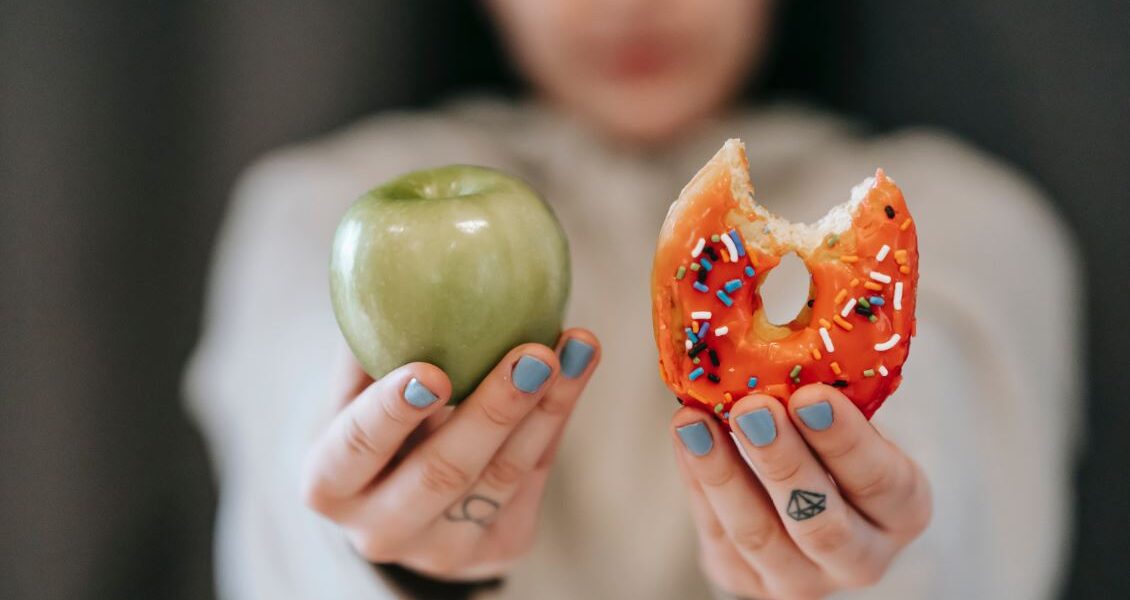 Mujer sosteniendo a cámara una manzana verde en una mano y una dona en la otra.