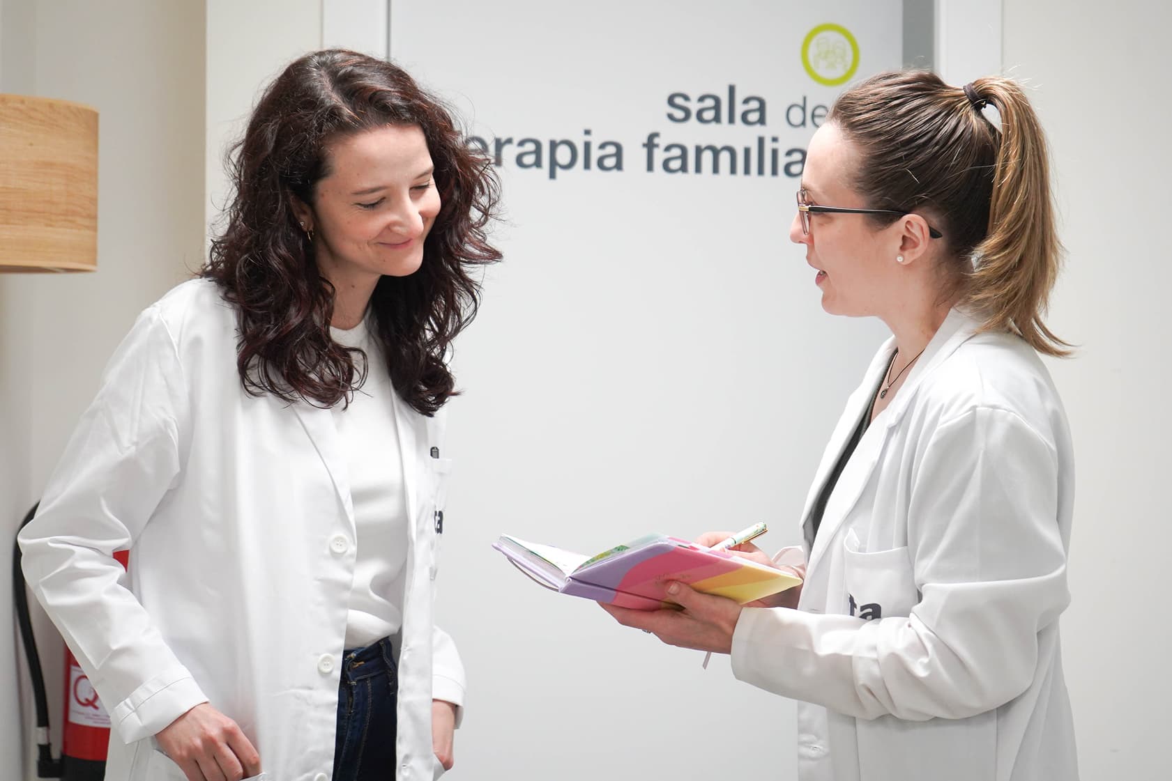 Una profesional de la salud en bata sosteniendo un libro mientras conversa con otra
