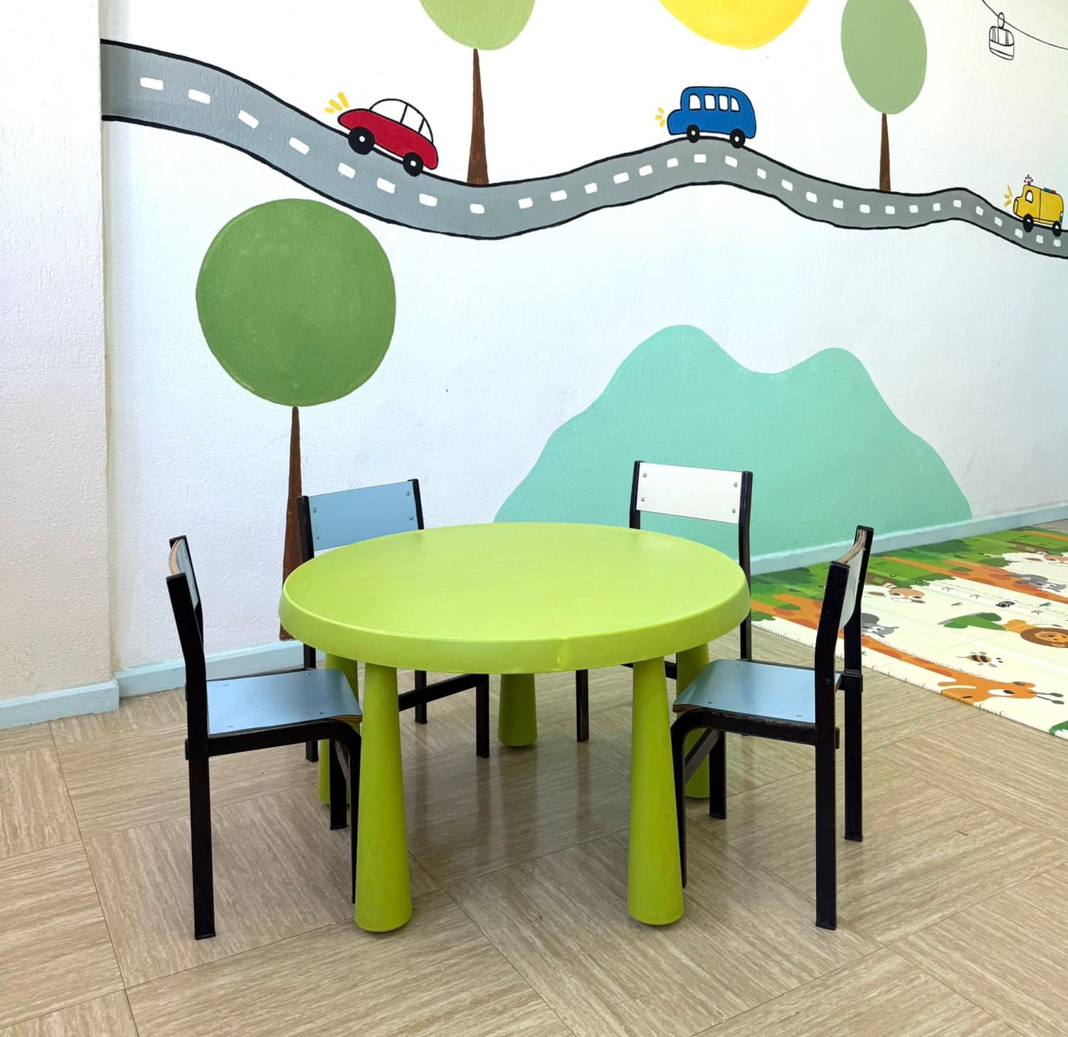 Mesa infantil verde con sillas frente a un mural de árboles y montañas